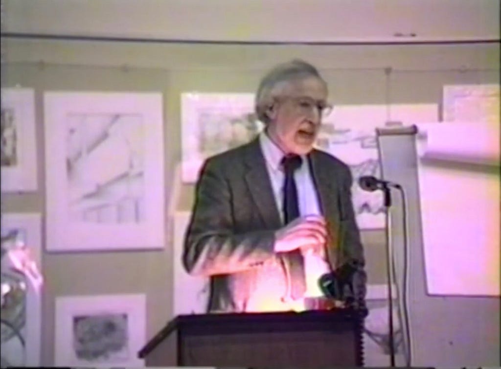 Donald Schön in a suit and tie speaks at a podium with a microphone. He has white hair and glasses, and is gesturing with one hand while holding papers in the other. Behind him are framed sketches or drawings on the wall, and a flip chart is visible to the side. The image has a grainy, vintage quality.