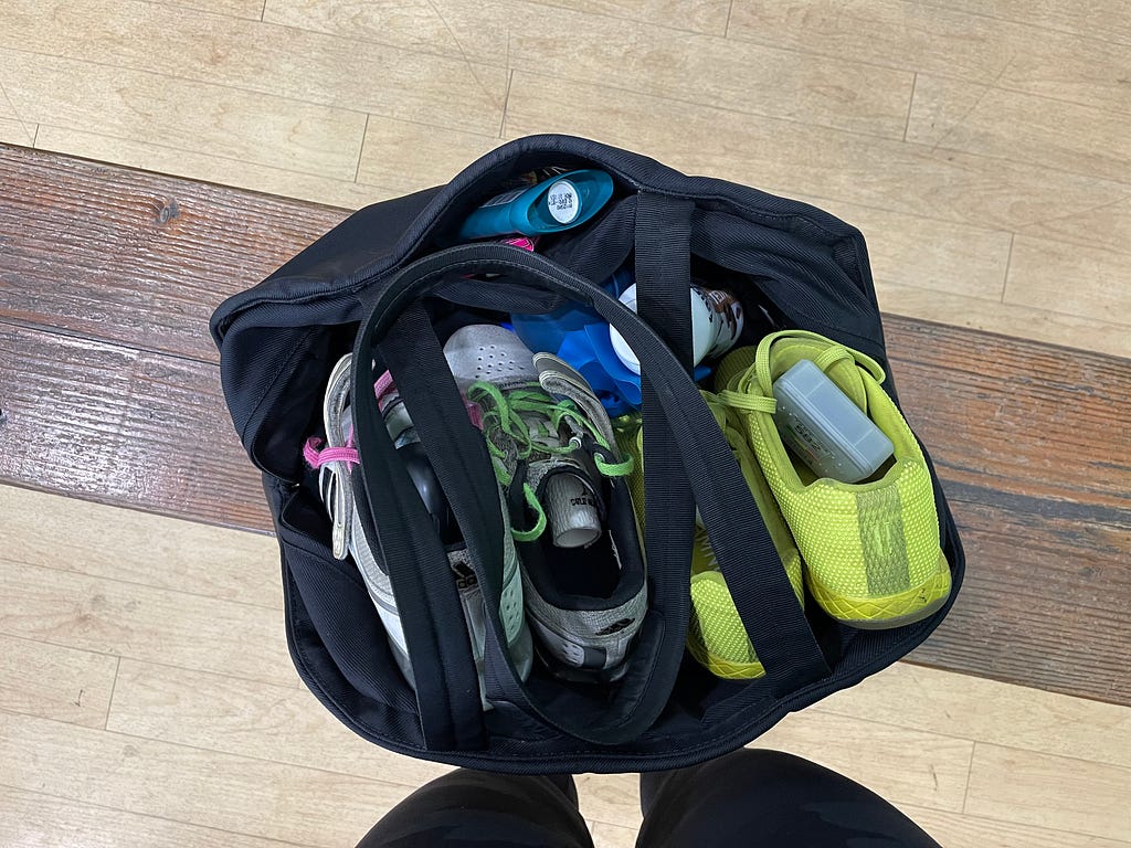 The writer’s gym bag, carefully filled with the necessary items, pictured from above as it sits open on a wooden bench inside the gym. Visible items include weightlifting shoes, CrossFit shoes, deodorant, a pack of gum, physical therapy bands, lifting thumb tape, an Aranet CO2 monitor, and a fairlife protein shake.