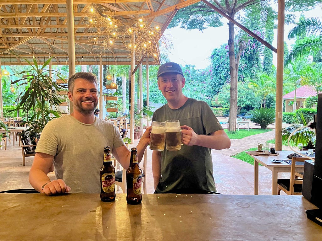 The author, pictured at right, toasts with a fellow climber holding mugs of beer with the bottles sitting on the bar and a resort-like setting in the background.