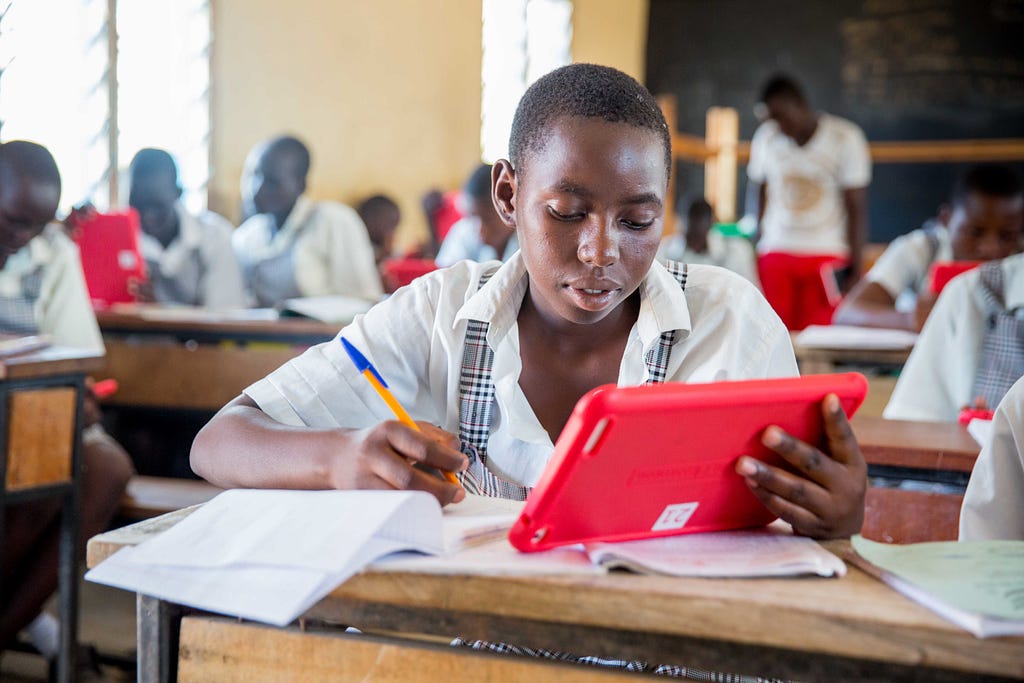 a girl learner sits at a desk with pen, notebook and a tablet in her hands