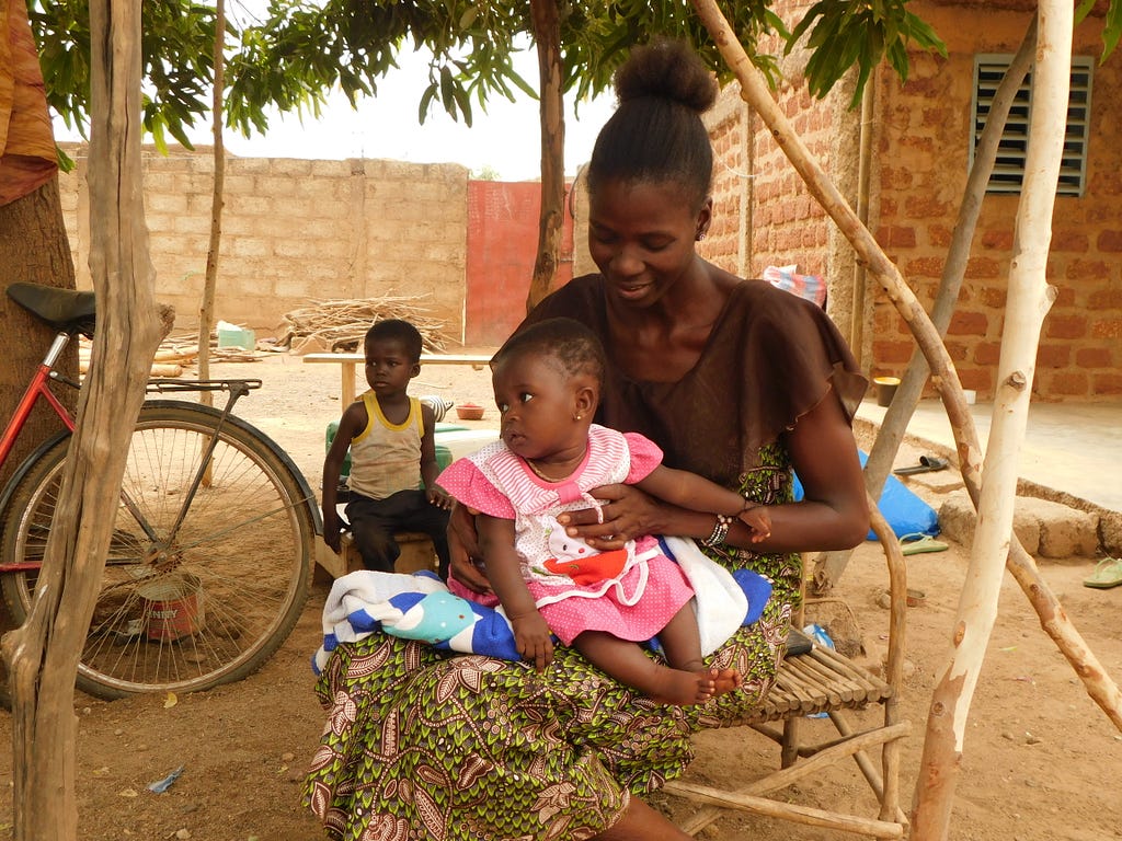 A young mother sits on a chair outside with a six-month-old girl on her lap dressed in pink.