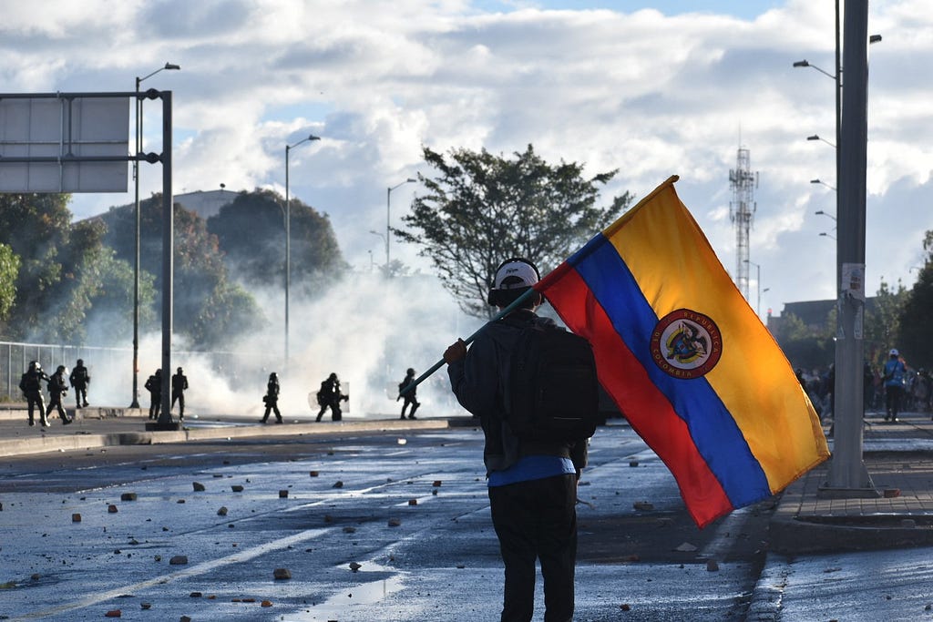 A protester holding a Colombian flag watches on as ESMAD riot police advance on protesters near Portal de Las Americas, Bogota, on Tuesday. Photo: Joshua Collins.