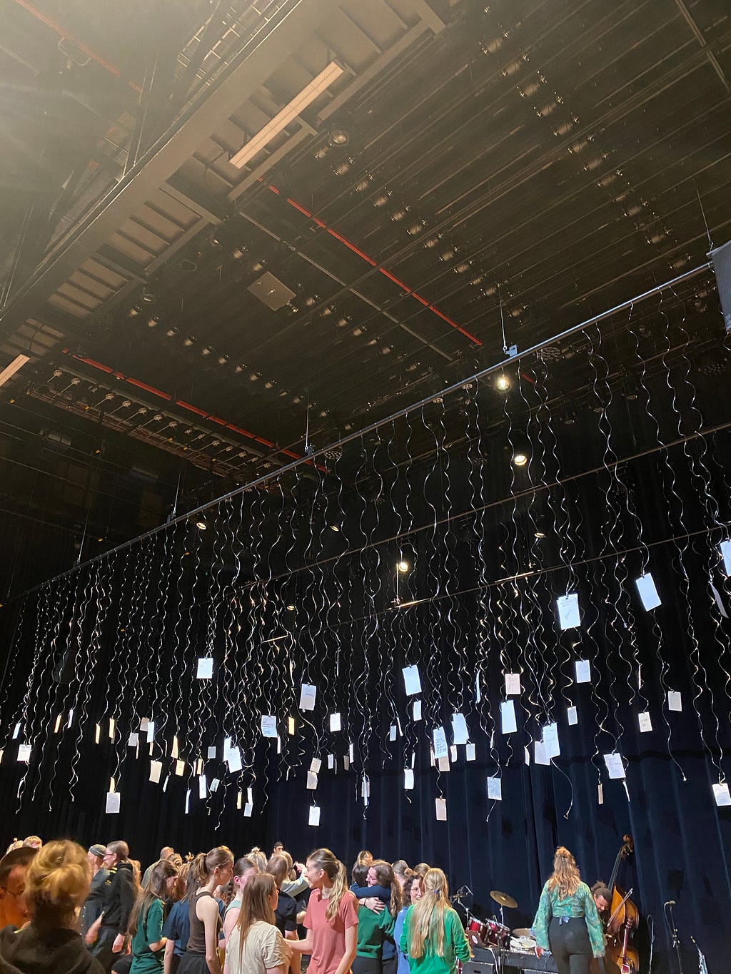 A theatre space with pieces of paper hanging from the top on strings and people interacting with each other underneath, as well as some musical instruments in the background.