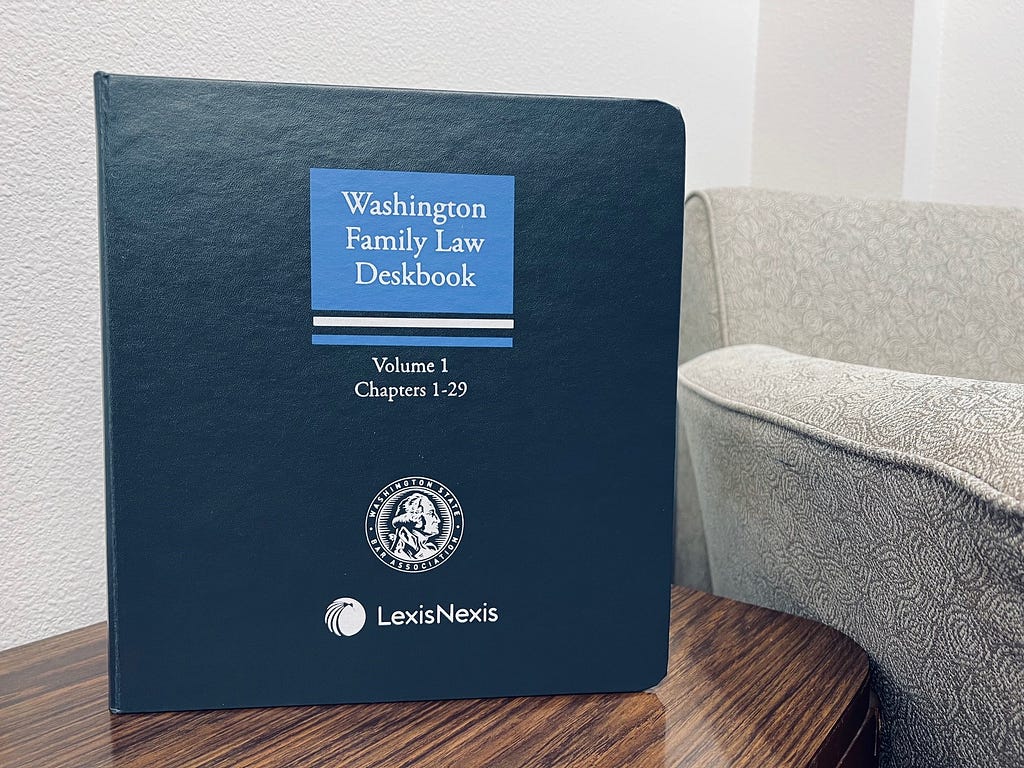 A blue book is standing upright on a piece of furniture next to an upholstered reading chair. The title of the book is Washington Family Law Deskbook.