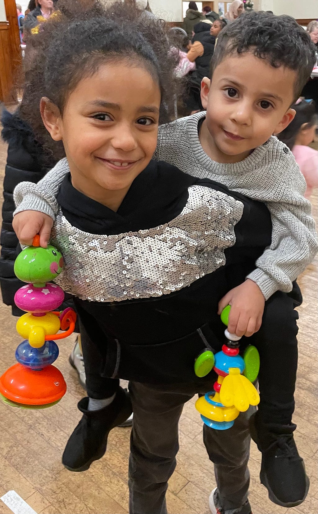 An image of two children, one about 7 and one about 4. The four year old is wrapped around the seven year old and holding toys. Both are smiling.