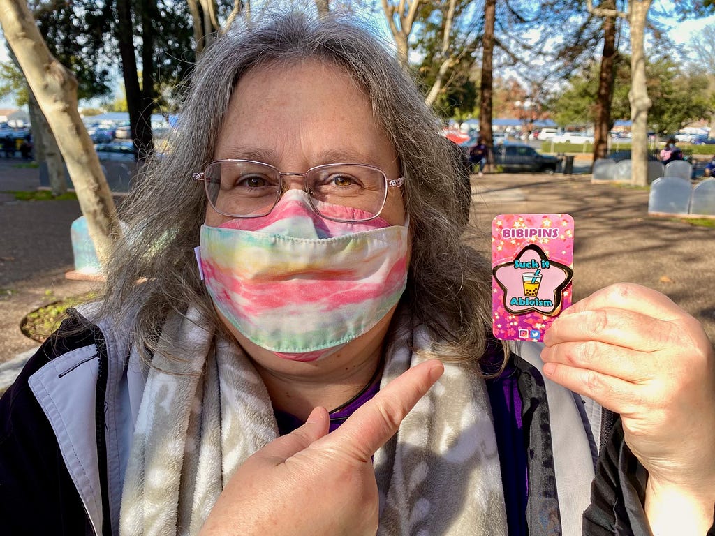 Michele Mashburn, a white woman with graying hair and glasses, wearing a mask pointing at a “Suck it Ableism” pin. There are trees behind her.