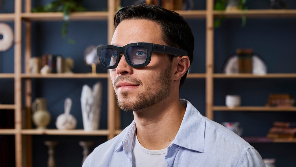 A man wearing Meta’s Orion AR glasses in a modern interior, with a neutral expression, suggesting casual integration of augmented technology into daily life.