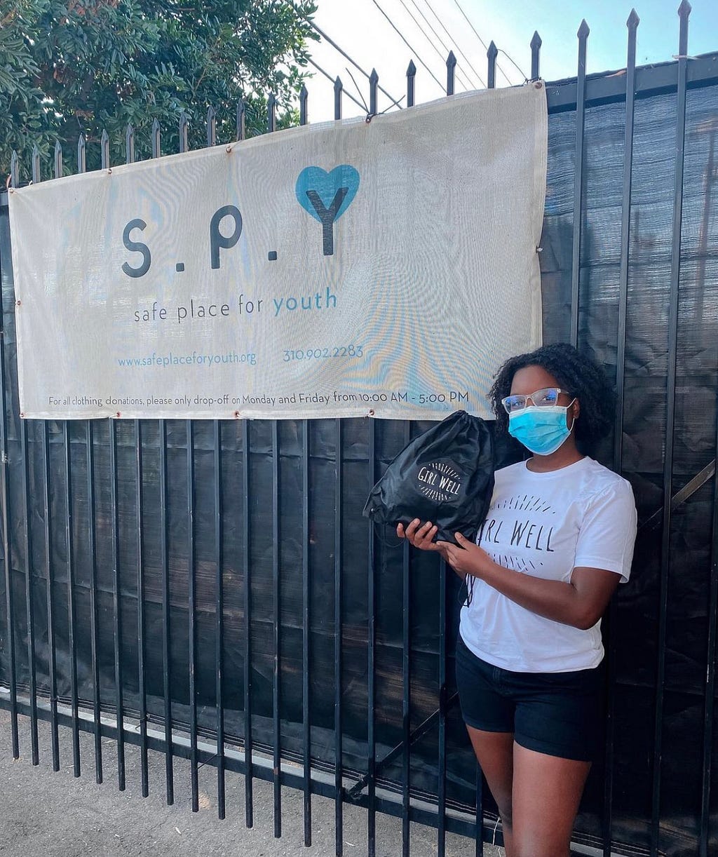 Young girl in glasses and a white “Girl Well” shirt posing with one of her kits in front of the entrance of “safe place for youth.”