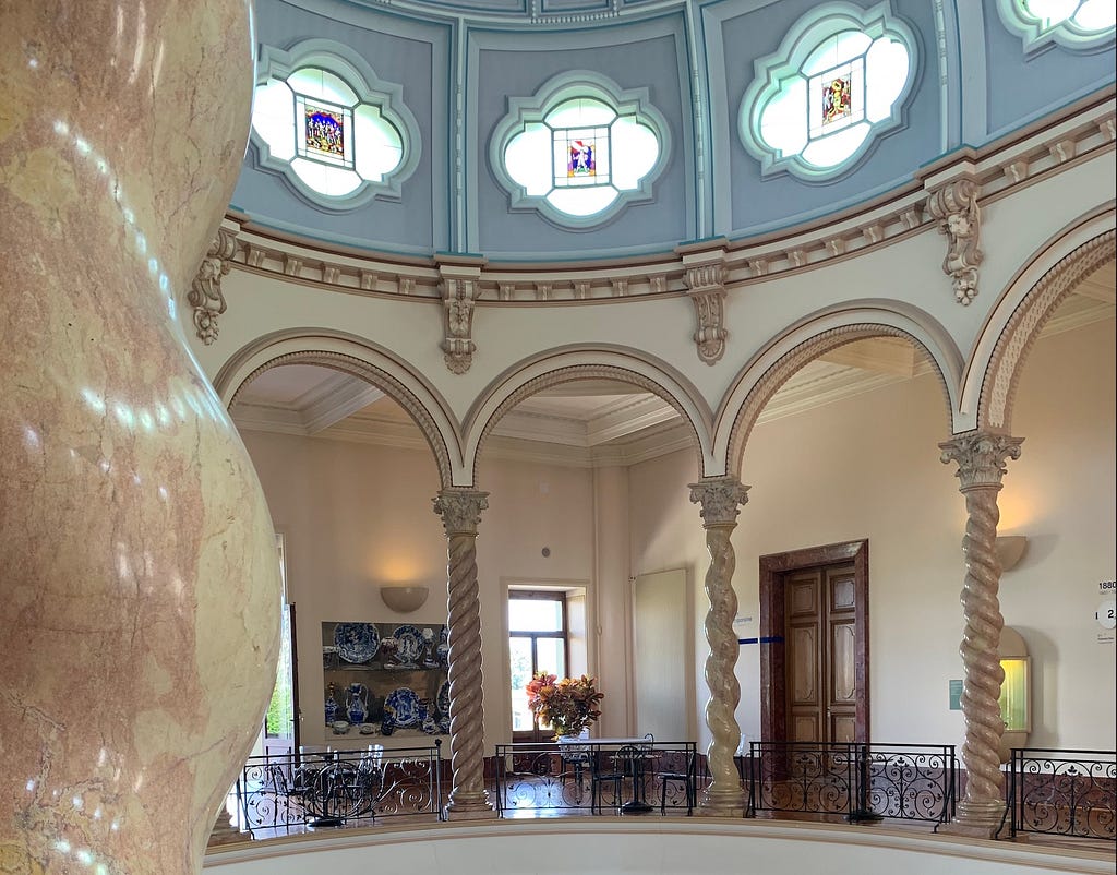Interior of Musée Ariana, showcasing a grand marble column in the foreground, elegant arches, a balcony with intricate ironwork, and a domed ceiling with stained glass windows.