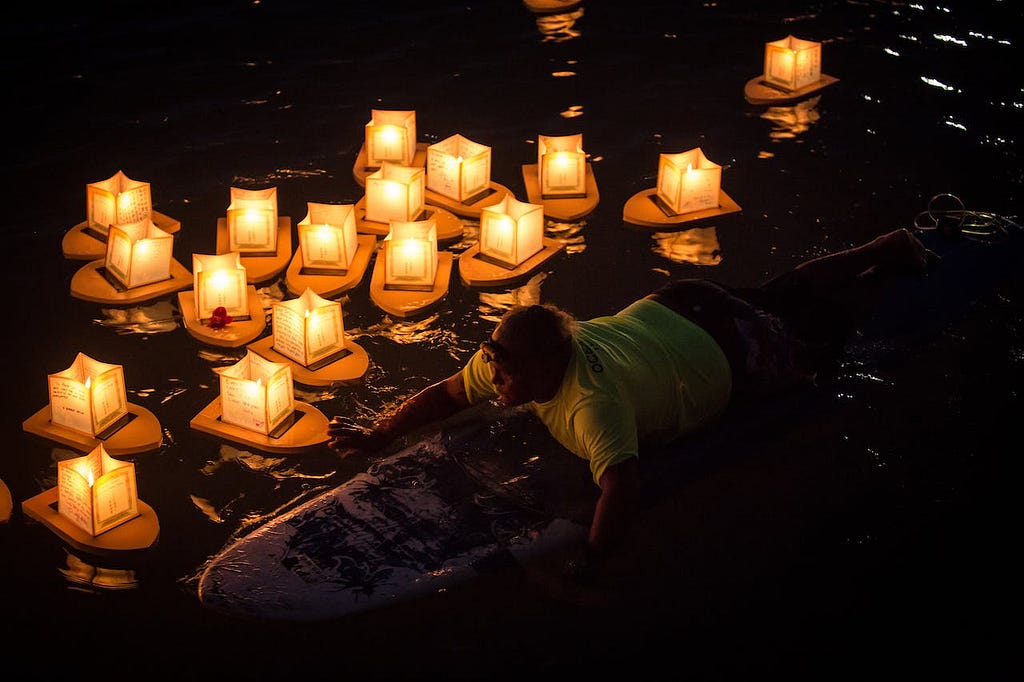 A man is lying on the ground next to a lake or river and floating a paper lantern. There are rows of lanterns already floating on the water.