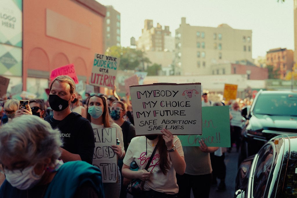 A group of people protesting for abortion rights and reproductive justice. They are wearing face masks and many are holding protest signs. A more prominent sign reads: My Body My Choice My Future. Safe abortion saves lives.