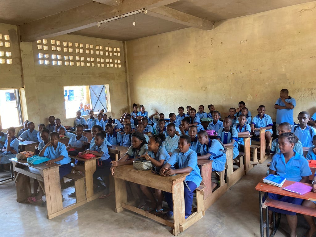 Group of young students, both boys and girls, wearing blue school uniforms, in a classroom, sitting at desks.