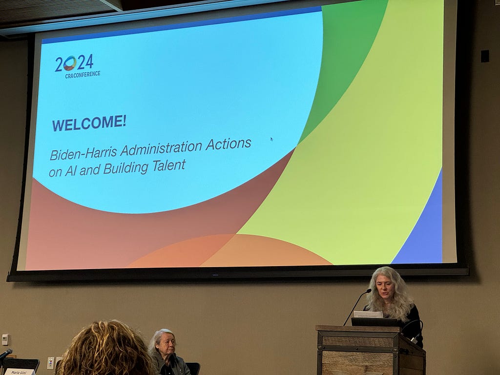 Deirdra on stage in front of a podium with long grey hair looking down at notes in front of a large slide that says “WELCOME! Biden-Harris administration actions on AI and building talent.”