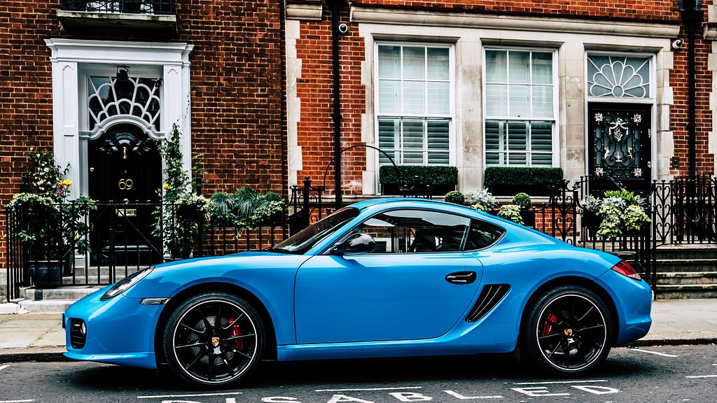 A light blue Porsche in front of a fancy home.
