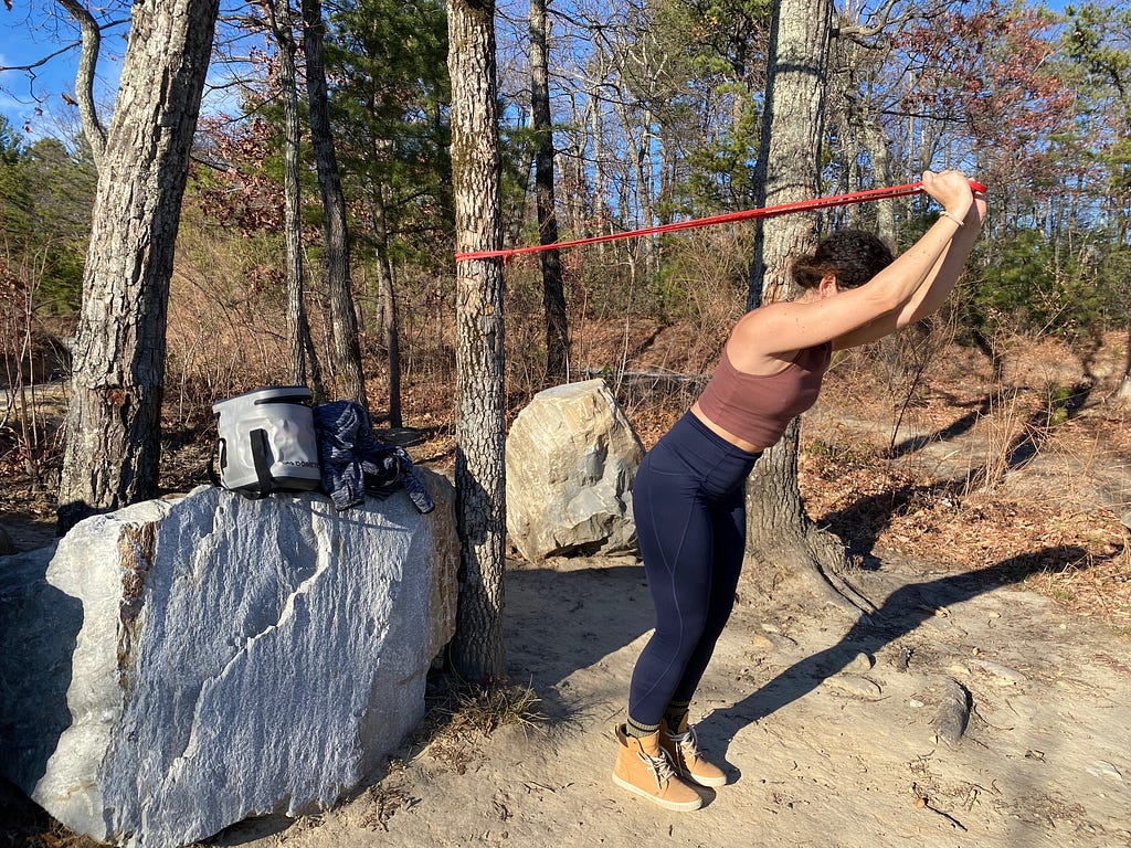 A woman performing triceps extension exercise with a red resistance band looped around a tree