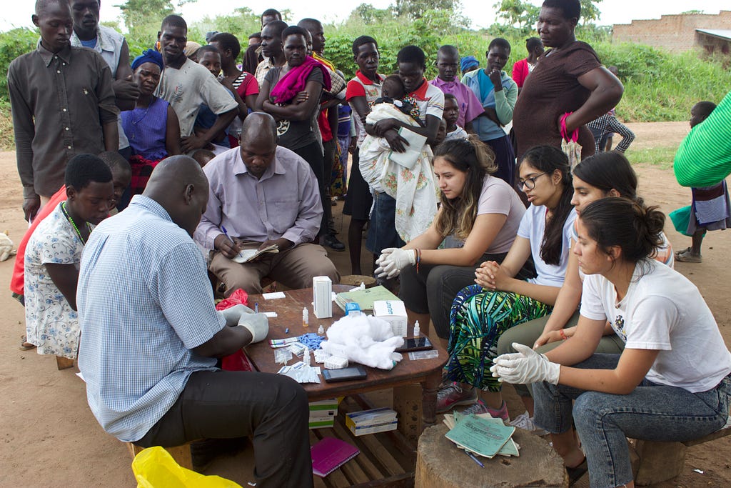 A group of people are gathered around a wooden table outside with medical supplies on it. On the left side of the table are staff members of GWED-G, and students are on the right side of the table observing and learning from the staff members. In the background, a group of people look on.