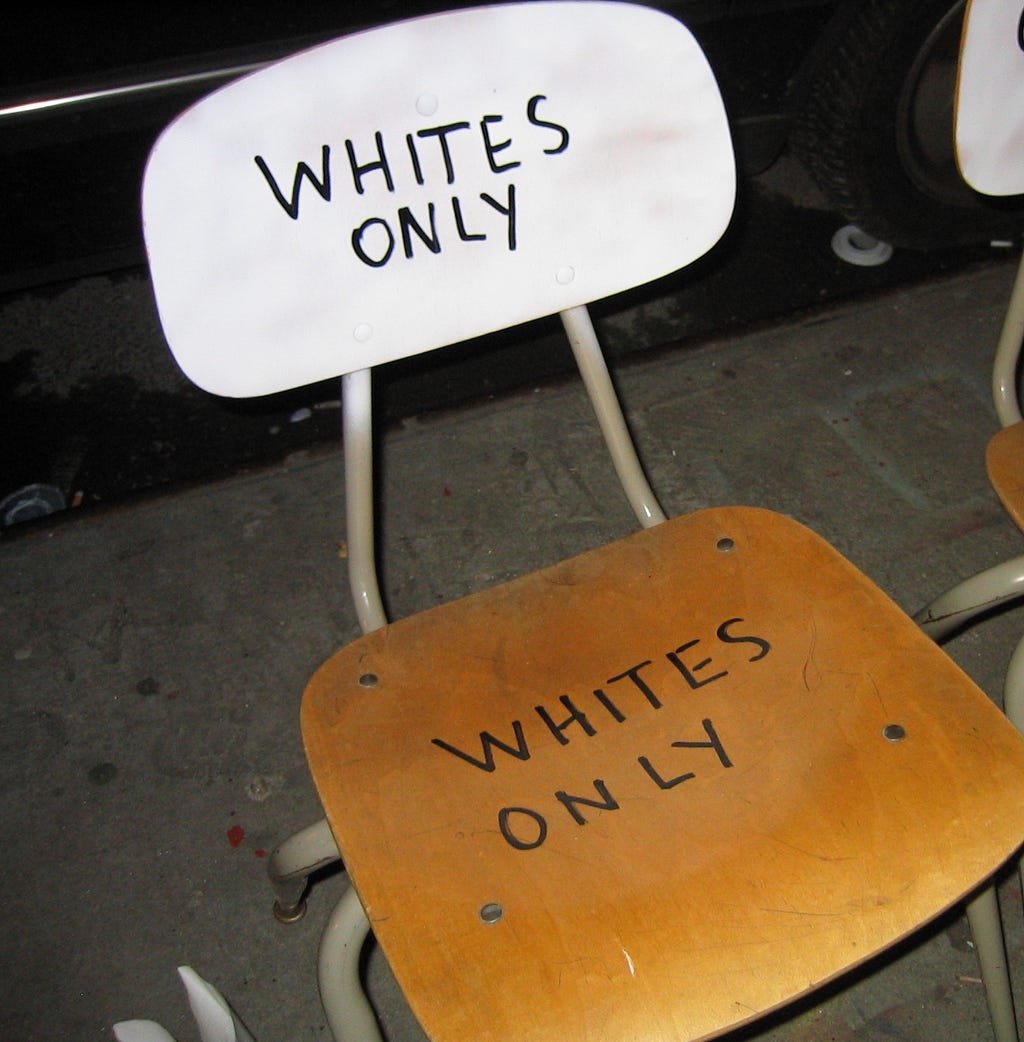 1950s elementary school wood chair with a white chair back and natural wood seat. On both surfaces, the words “Whites Only” are hand-written in thin, black marker.