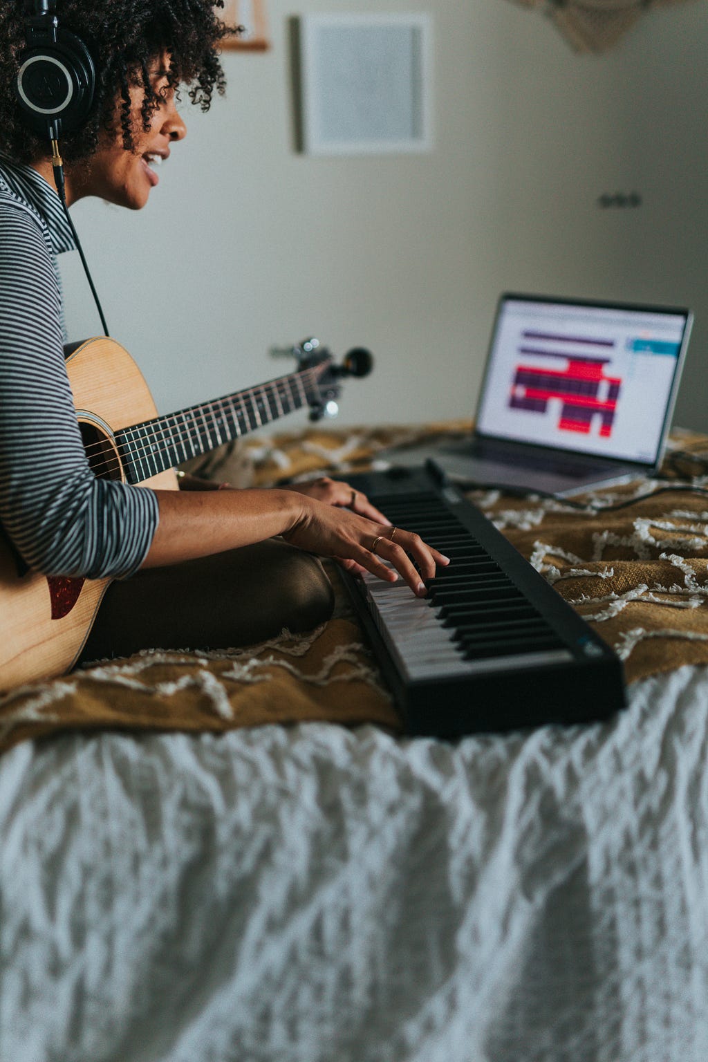 A young musician works on a keyboard and a guitar in their bedroom.