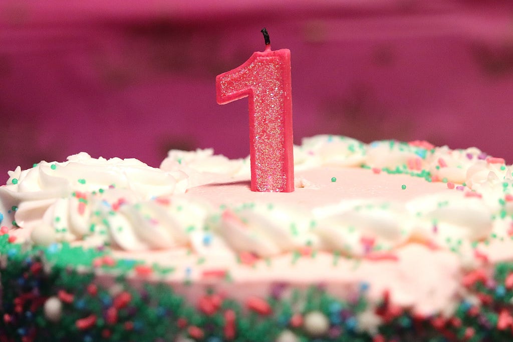 An unlit pink candle in the shape of the number one is seen on the top-half of a round cake covered with white icing and green and pink sprinkles, against a fuchsia background.