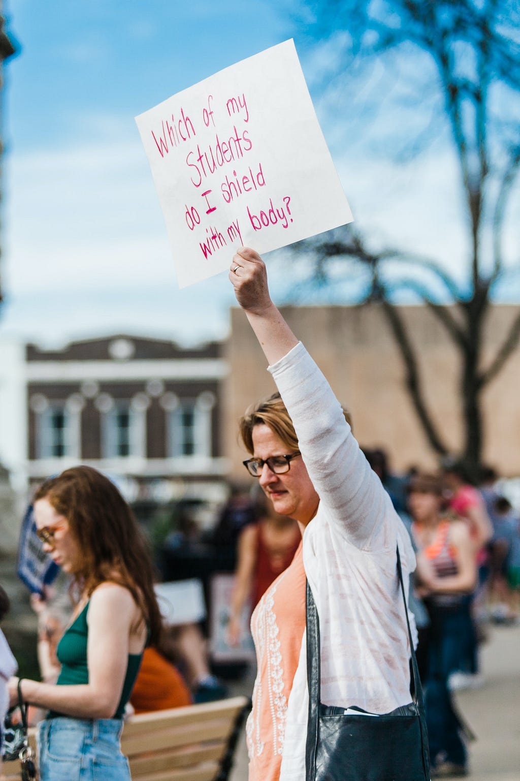 A woman holds up a sign that reads “Which of My Students Do I Shield With My Body?” Photo credit Heather Mount/Unsplash