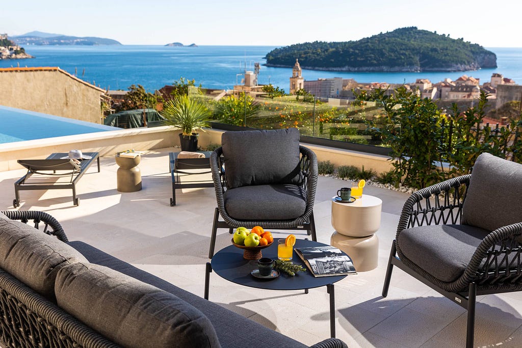 A scenic outdoor terrace overlooking a stunning coastal landscape. The terrace features modern outdoor furniture, including comfortable cushioned chairs and sofas arranged around a small round table with fruits and beverages. Nearby are lounge chairs positioned to enjoy the sun. The area is adorned with potted plants and vibrant flowers. In the background, a clear blue sea stretches out towards distant islands and a picturesque town with historic buildings and a church tower.
