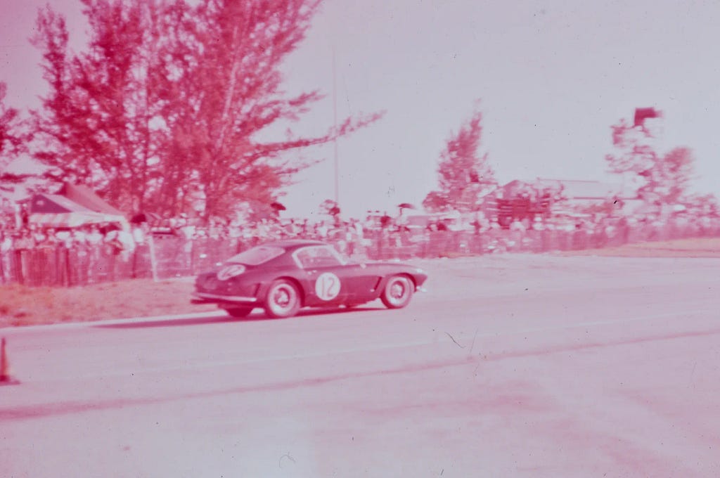 A faded color photo shows a black Ferrari with the number 12 on its side next to a fenced-off crowd of onlookers.