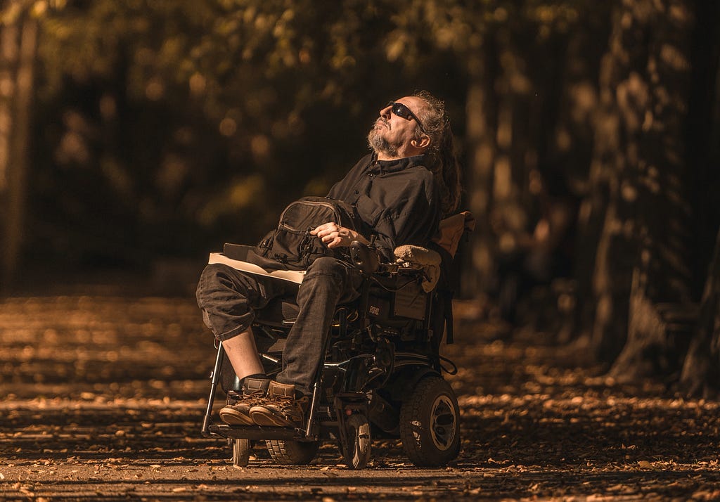 a man with sunglasses sitting in a wheelchair in a long alley with trees