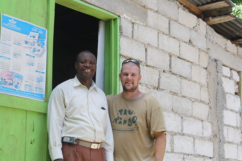 Black man in button down shirt standing next to sweaty, bald white man in front of school building in Haiti.