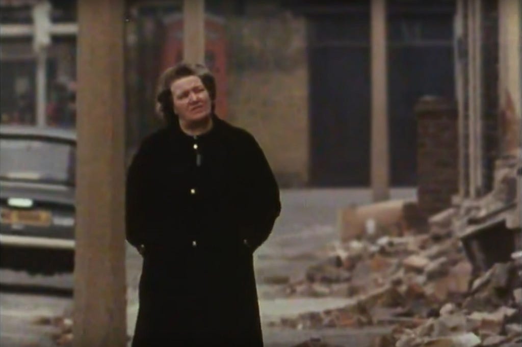 Still from Where the Houses Used to Be (1969). A woman in a heavy overcoat walks by a row of terraced houses currently being demolished.