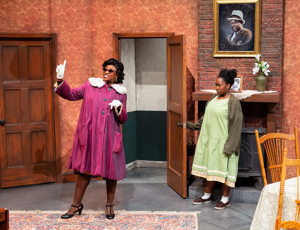 A woman in a pink 1950s coat and sunglasses gesticulates in an open door to an apartment while a young woman in a green 1950s dress looks on in confusion