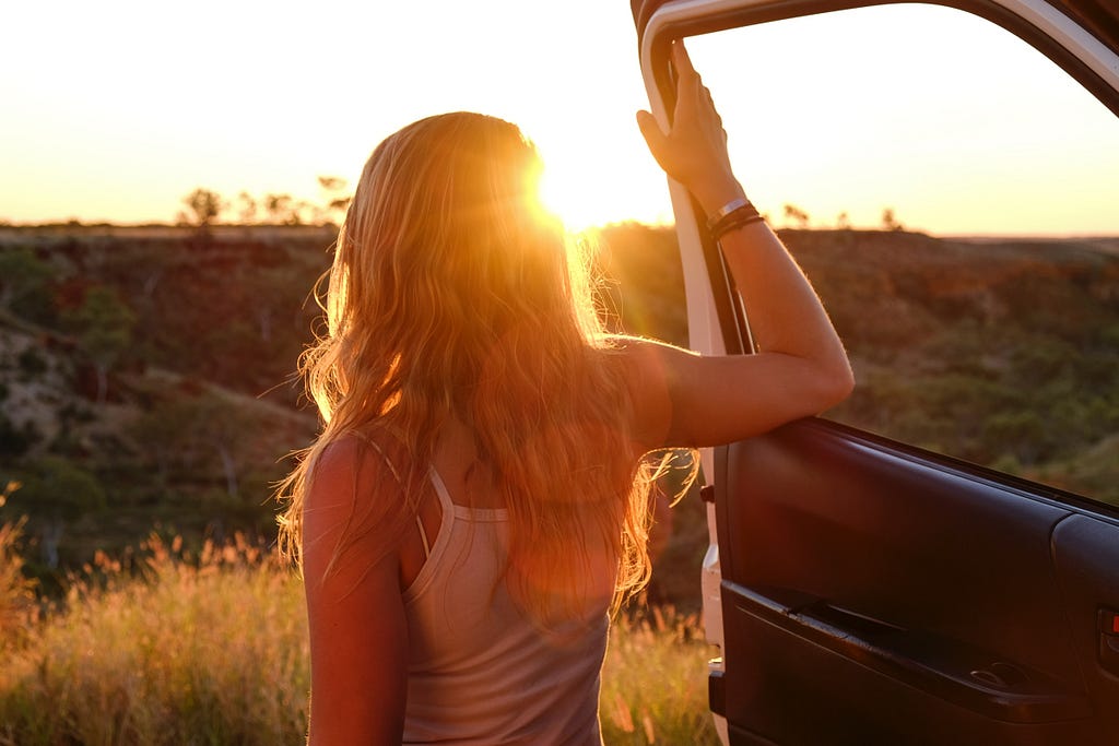 A blonde woman in a tank top stands outside a vehicle with her arm resting against the car door, facing the sunset