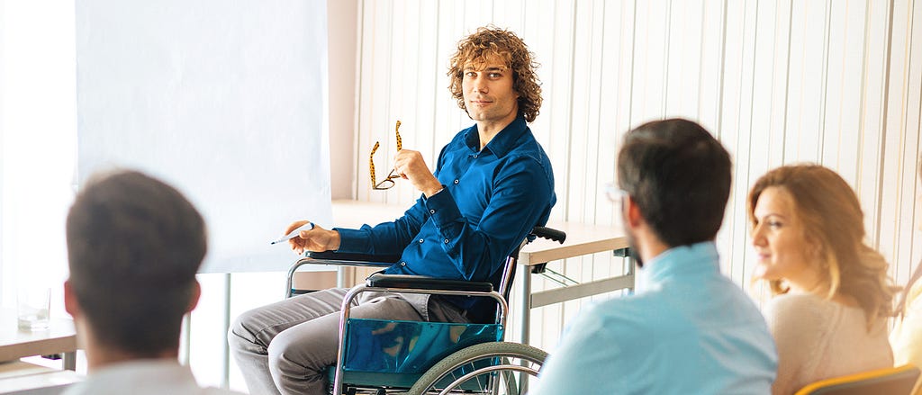 In a professional training session, a curly-haired male instructor sits in a wheelchair in front of a flipchart.