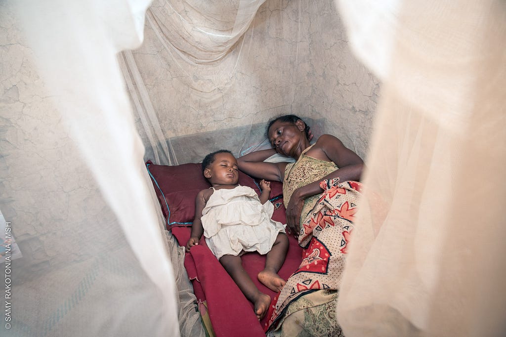 A woman lies in bed next to a sleeping baby under a mosquito net