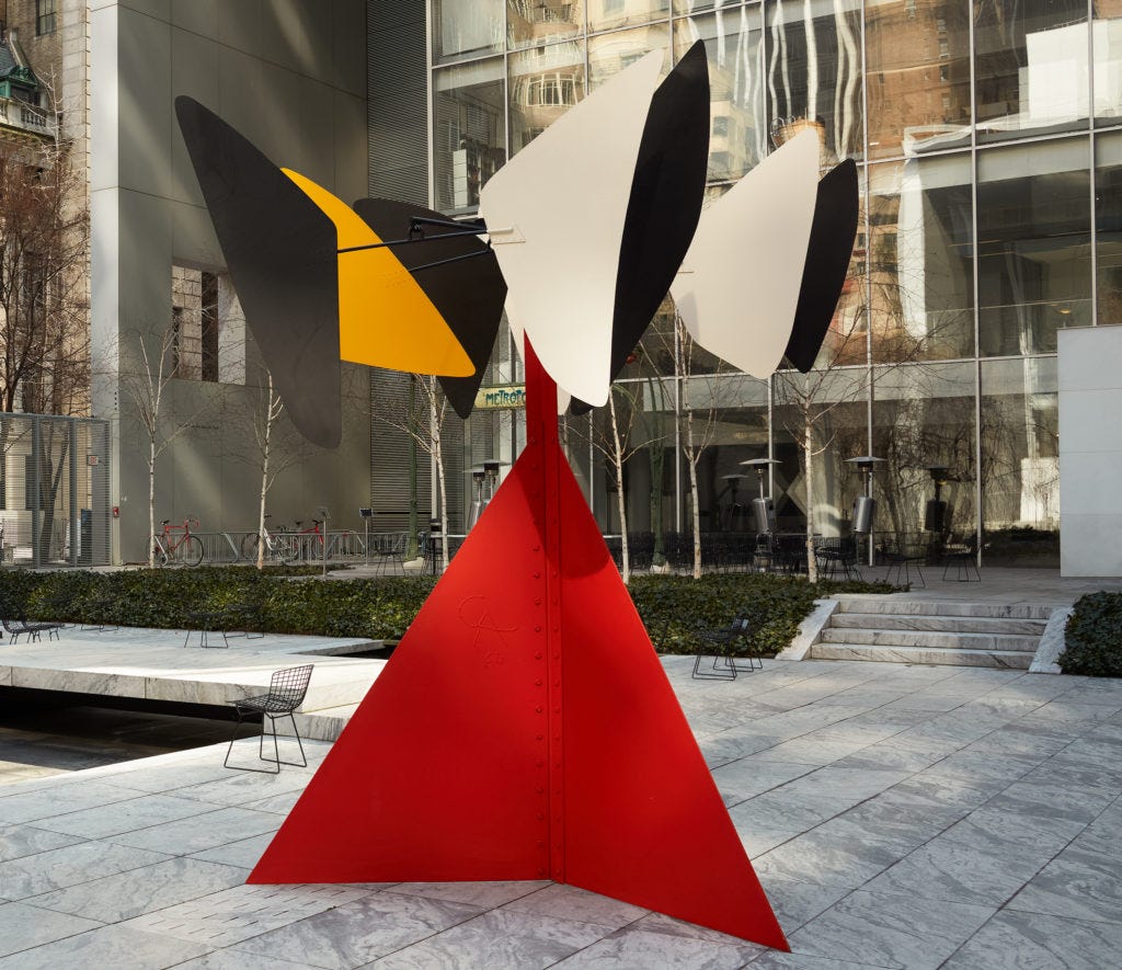 In a paved plaza, a steeply angled red metal base, several feet tall, and triangular in shape, sits like a pyramid with the point at the top, to support a rotating carousel of six or seven smaller, more smoothly rounded triangular metal panels.