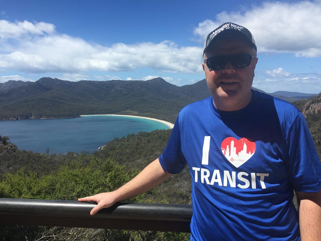 A man wearing a blue shirt that reads “I heart Transit” stands in front a blue body of water.