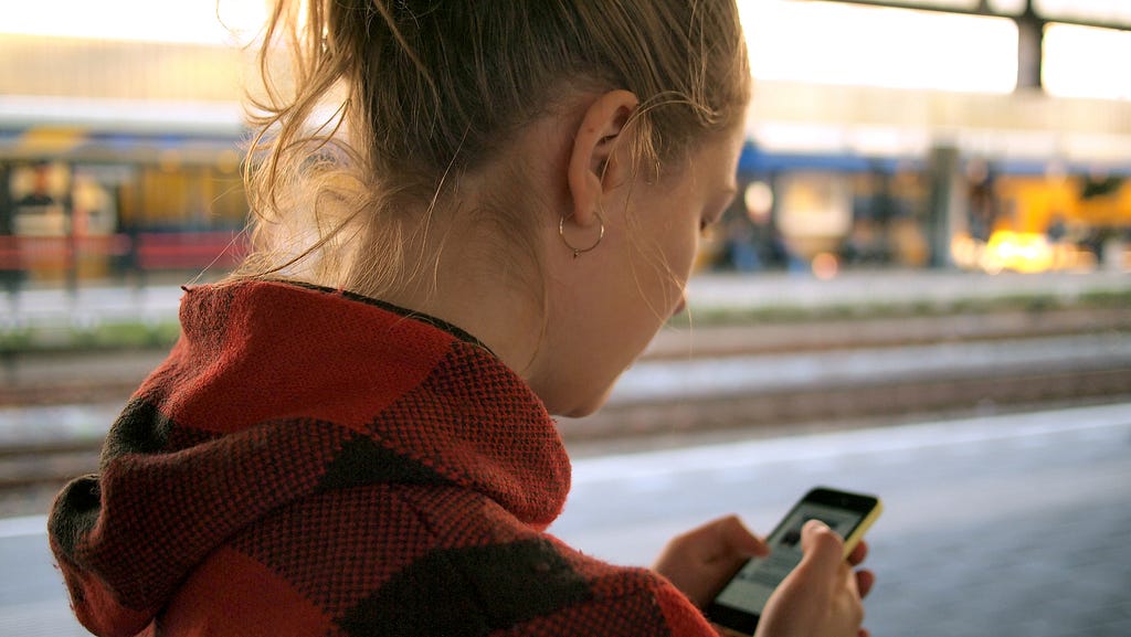A photograph of a woman standing on a train platform using her smartphone