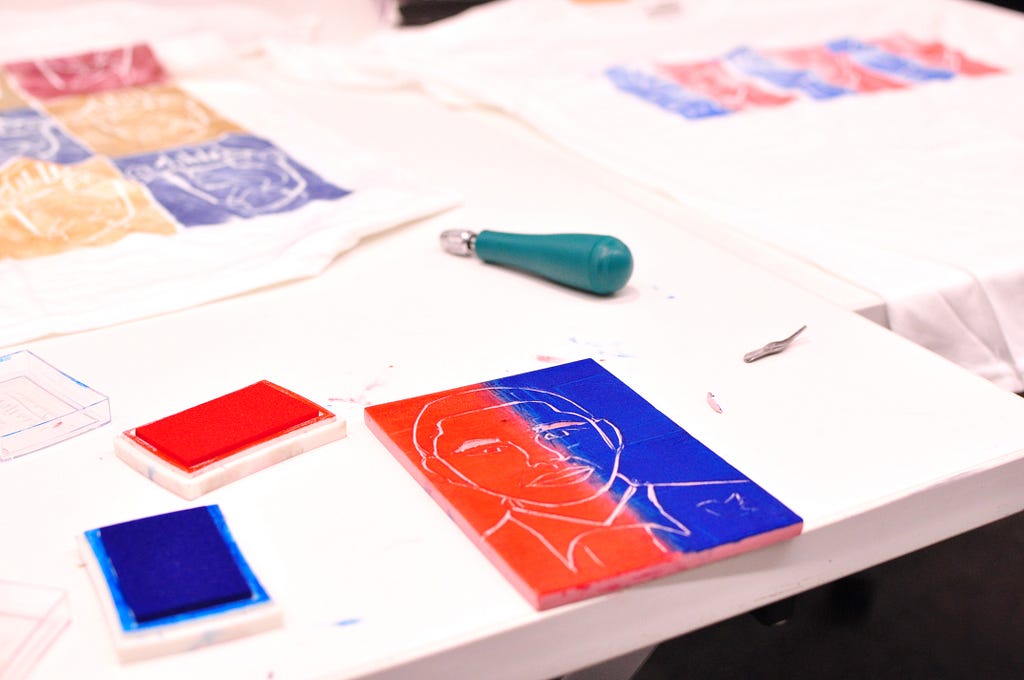 In the foreground of a photo, red and blue ink pads are next to a rubber block stamp of President Obama, which has been covered half in red ink and half in blue ink. In the background, a carving tool lays next to 2 t-shirts that have been stamped.