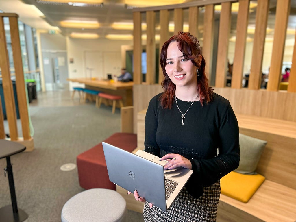 A photo of Orla Dunlop,DevOps graduate at the Department for Work and Pensions holding a laptop smiling at a office setting.