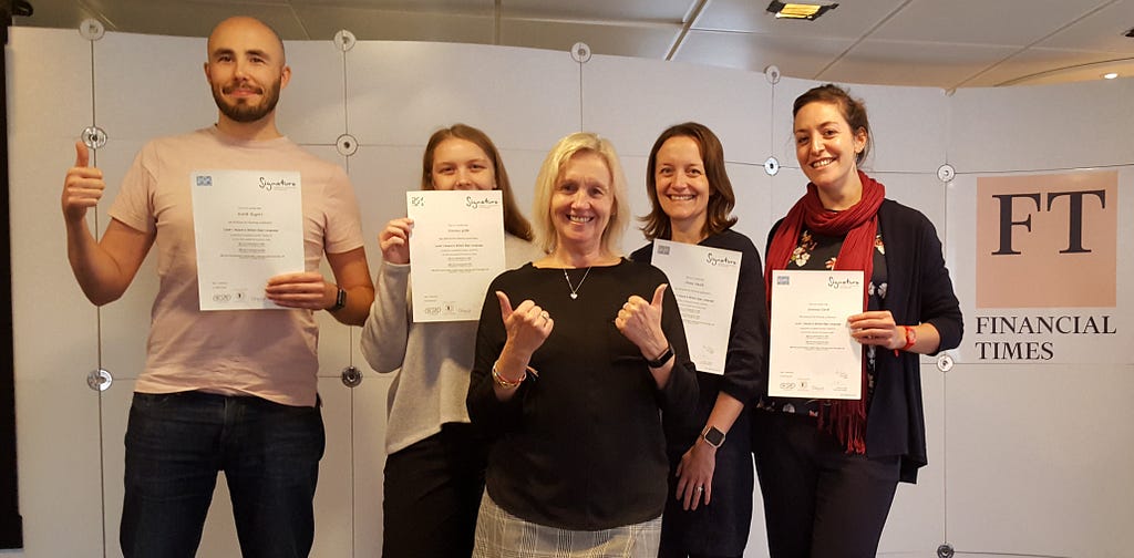 Four students display their certificates for passing the British Sign Language 101 examination and are accompanied by their teacher who is giving a double thumbs up.