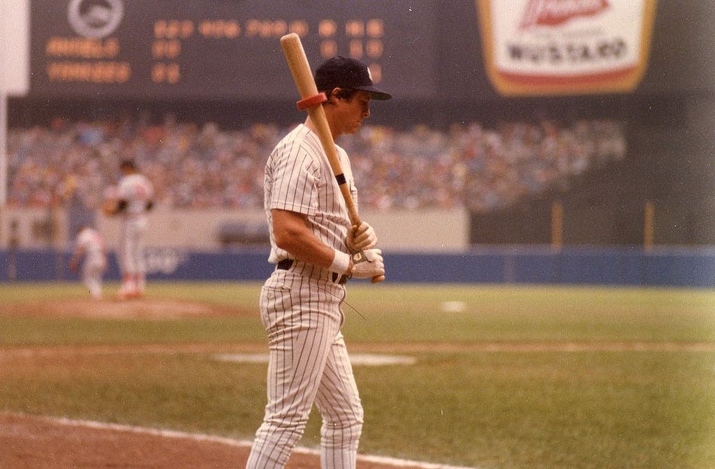 Bobby Murcer holding a bat with a weighted red “donut” on the barrel. He is standing, in profile, looking down as he waits his turn to bat.