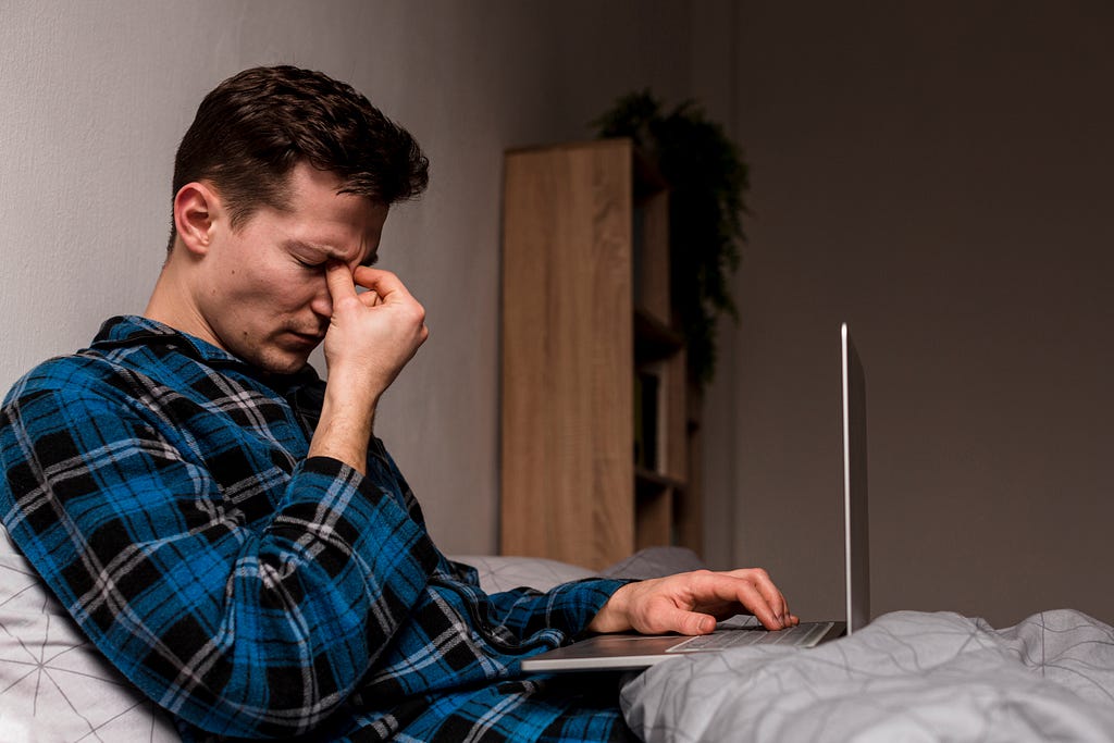 An exhausted young man lies in bed, surrounded by dim lighting, laptop open in front of him.