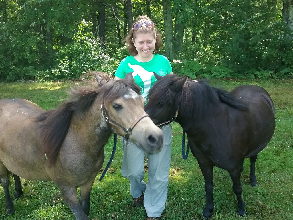 a woman smiles and stands with two small horses