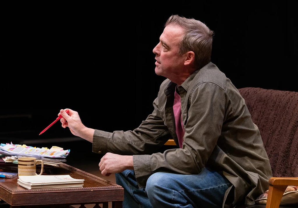 A man in a brown shirt and jeans sits on a couch holding a red pen. The table in front of him has a neat stack of notebooks and a messy stack of paper.