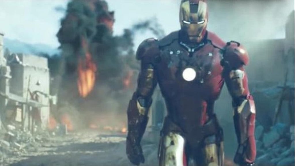 Iron Man’s gold-and-red suit, on the right side of the image, appears to be walking away from an explosion (orange flames and black smoke) in a sandy, barren area.