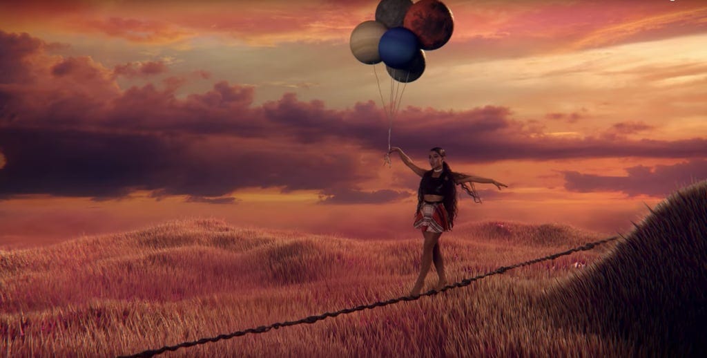 An image of Ariana Grande walking a tightrope, balancing balloons. Image from music video “God is a Woman”.