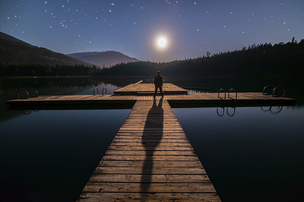man standing at the end of a dock, surrounded by water, staring at the night sky and contemplating