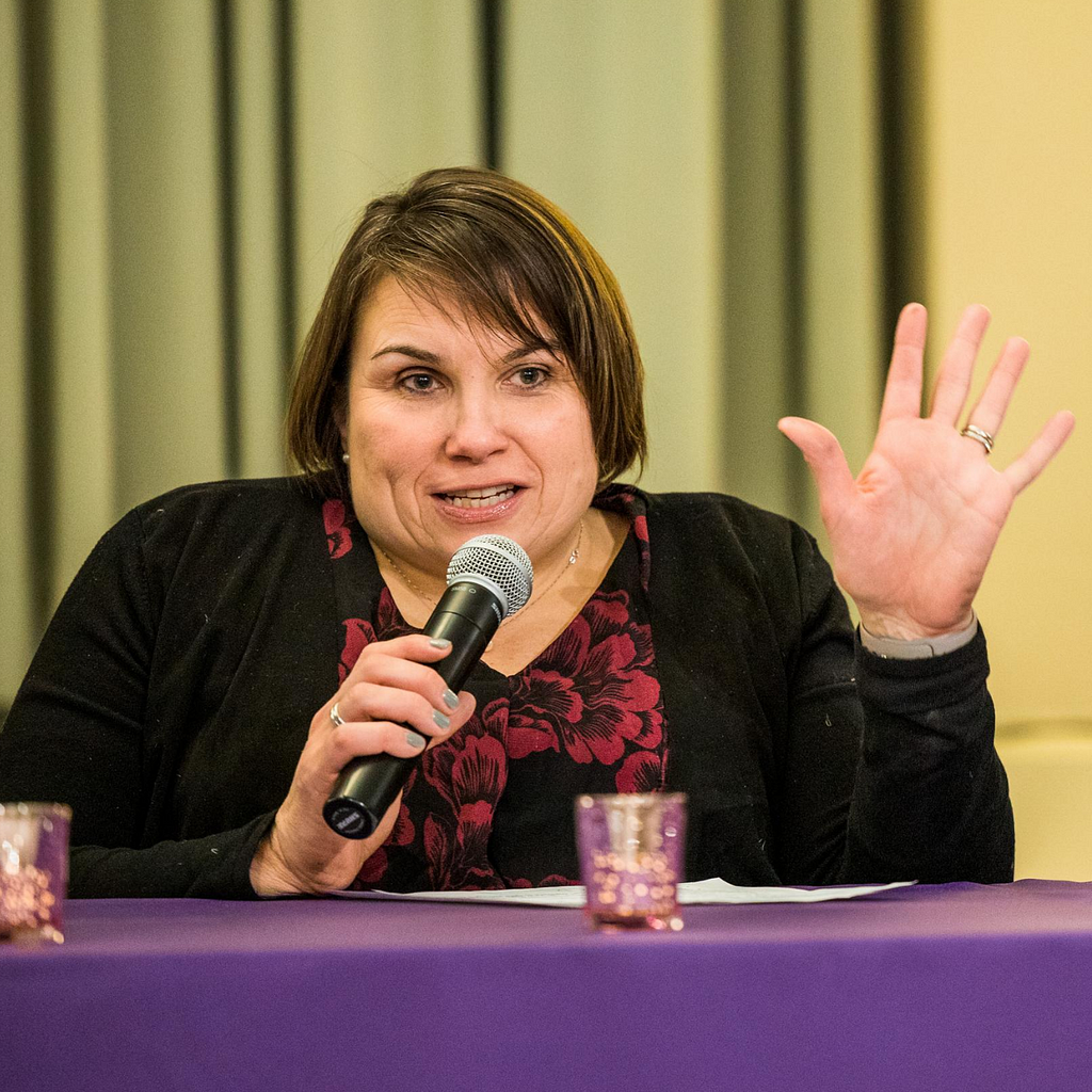 Karen speaks into a microphone, her left hand gesturing outward. Her hair is short, brown. Red floral blouse, black cardigan.