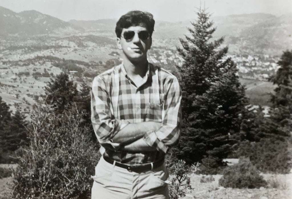 Man standing with arms crossed and sunglasses, standing in front of pine trees with mountains in the background.