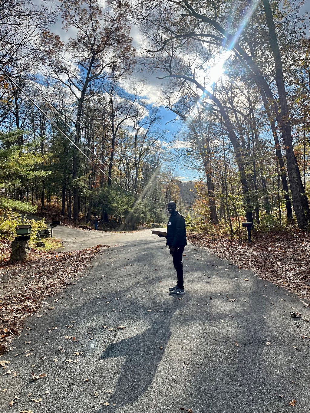 A man stands on a rural road holding a clipboard. He is surrounded by trees and autumn leaves, with the sun peering through gaps in the clouds.