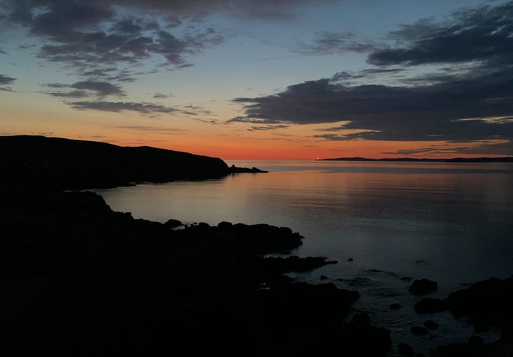The silhouette of scottish coast above the sea at sunset, with the light of a light house in the distance.
